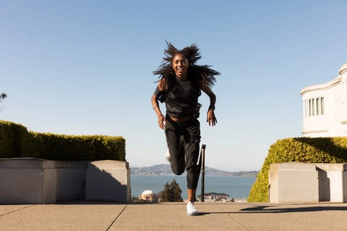 Athletic African-American woman running outdoors on a sunny day. She is wearing sportswear with ocean and city skyline in background.