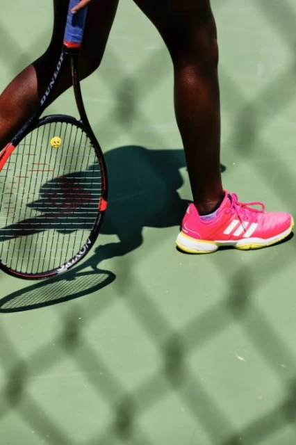 A tennis player holds a racket on a green court, with a close-up of the racket, pink sports shoe, and lower legs in bright daylight.