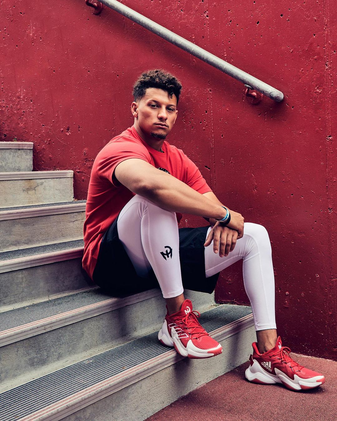 Man in athletic wear sits on concrete steps against a red wall, showcasing an athletic and casual demeanor in an urban environment.