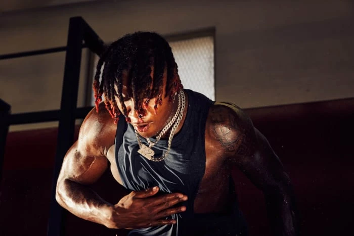 An adult male with dreadlocks and a gold chain in a tank top is in a gym setting, engaging in a workout routine.