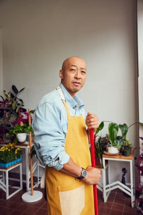 Adult male in a yellow apron stands indoors holding a red mop, surrounded by potted plants on tables and wearing a blue shirt.