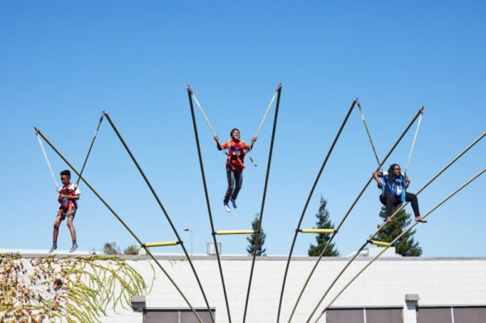 Three people enjoy a bungee trampoline experience on a sunny day with a clear blue sky. They are suspended mid-air by elastic cords.