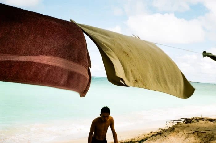 A young man walks along a sandy beach with towels hanging above on a clothesline. Turquoise ocean and a partly cloudy sky in the background.