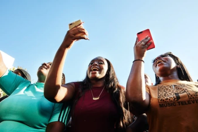 Three young Black women joyfully record a performance, holding up phones against a blue sky, wearing casual attire. Upward angle.