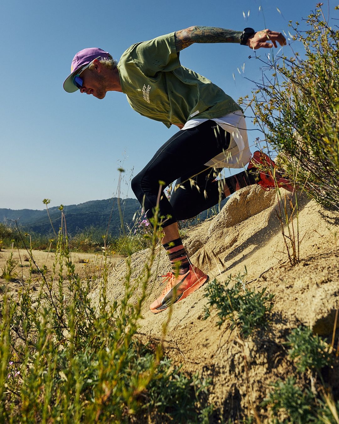 A man with tattoos runs downhill on a sandy trail in running gear, captured in an eye-level shot against a blue sky and mountain backdrop.