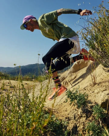 A man with tattoos runs downhill on a sandy trail in running gear, captured in an eye-level shot against a blue sky and mountain backdrop.