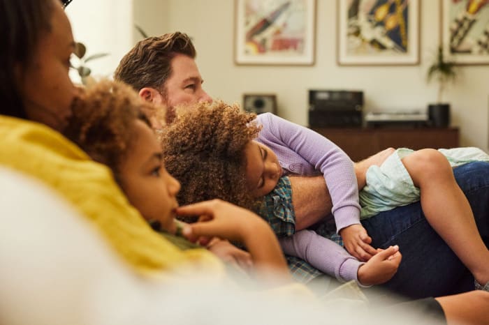 Family relaxing at home, two daughters resting on father while watching television in a brightly lit living room with neutral tones.