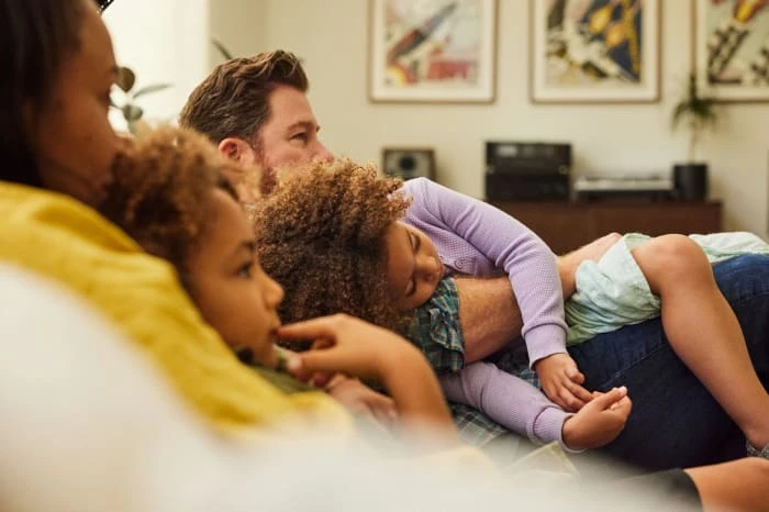 Family relaxing at home, two daughters resting on father while watching television in a brightly lit living room with neutral tones.