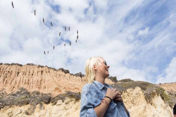 Woman smiles up at birds flying over a bluff and cloudy sky; she wears a denim shirt and bracelets in an outdoor, daytime shot.