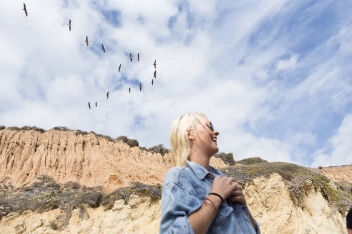 Woman smiles up at birds flying over a bluff and cloudy sky; she wears a denim shirt and bracelets in an outdoor, daytime shot.