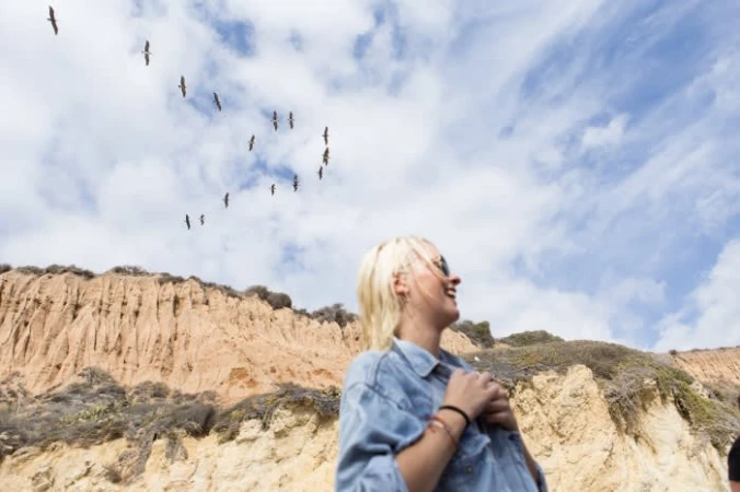 Woman smiles up at birds flying over a bluff and cloudy sky; she wears a denim shirt and bracelets in an outdoor, daytime shot.
