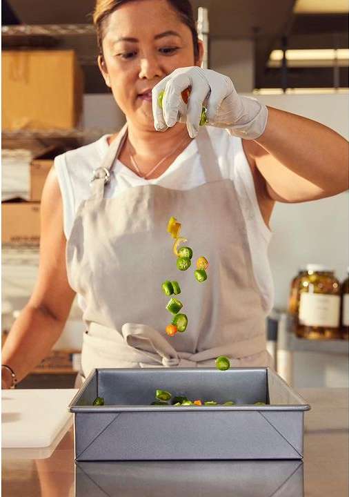 Woman in apron preparing green peppers in a kitchen, wearing gloves, chopping vegetables, and using a metal baking pan.