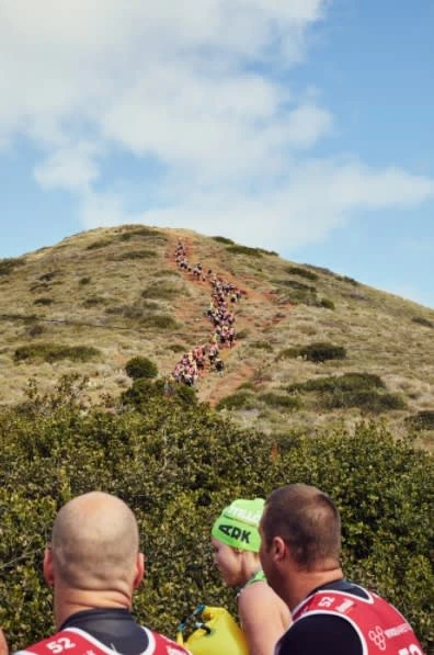 A group of athletes participates in a race up a grassy hill under a cloudy sky, as observed by people in athletic gear.