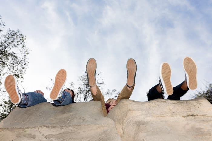 Three individuals relaxing outdoors with their feet up in canvas shoes, showcasing a carefree lifestyle against a cloudy sky.