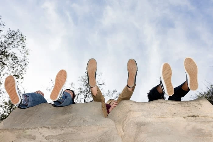 Three individuals relaxing outdoors with their feet up in canvas shoes, showcasing a carefree lifestyle against a cloudy sky.