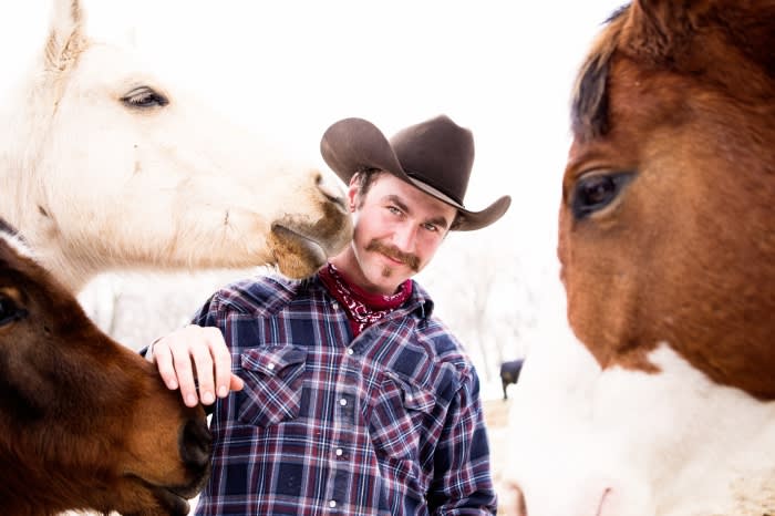 Portrait of a man wearing a cowboy hat with two horses. One light horse is near his head, and a brown horse is in the foreground.