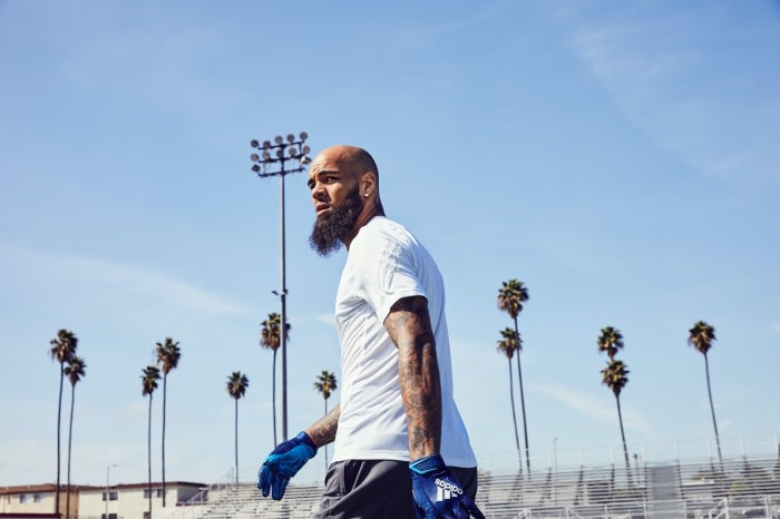 Man in white t-shirt and sport gloves gazes left on a track with stadium lights and palm trees under a clear blue sky.