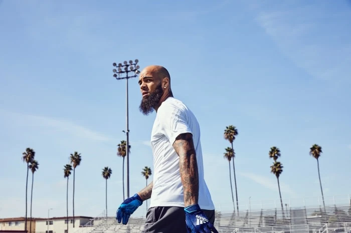 Man in white t-shirt and sport gloves gazes left on a track with stadium lights and palm trees under a clear blue sky.