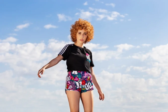 Young woman with curly red hair wearing a black t-shirt and colorful patterned shorts against a blue sky