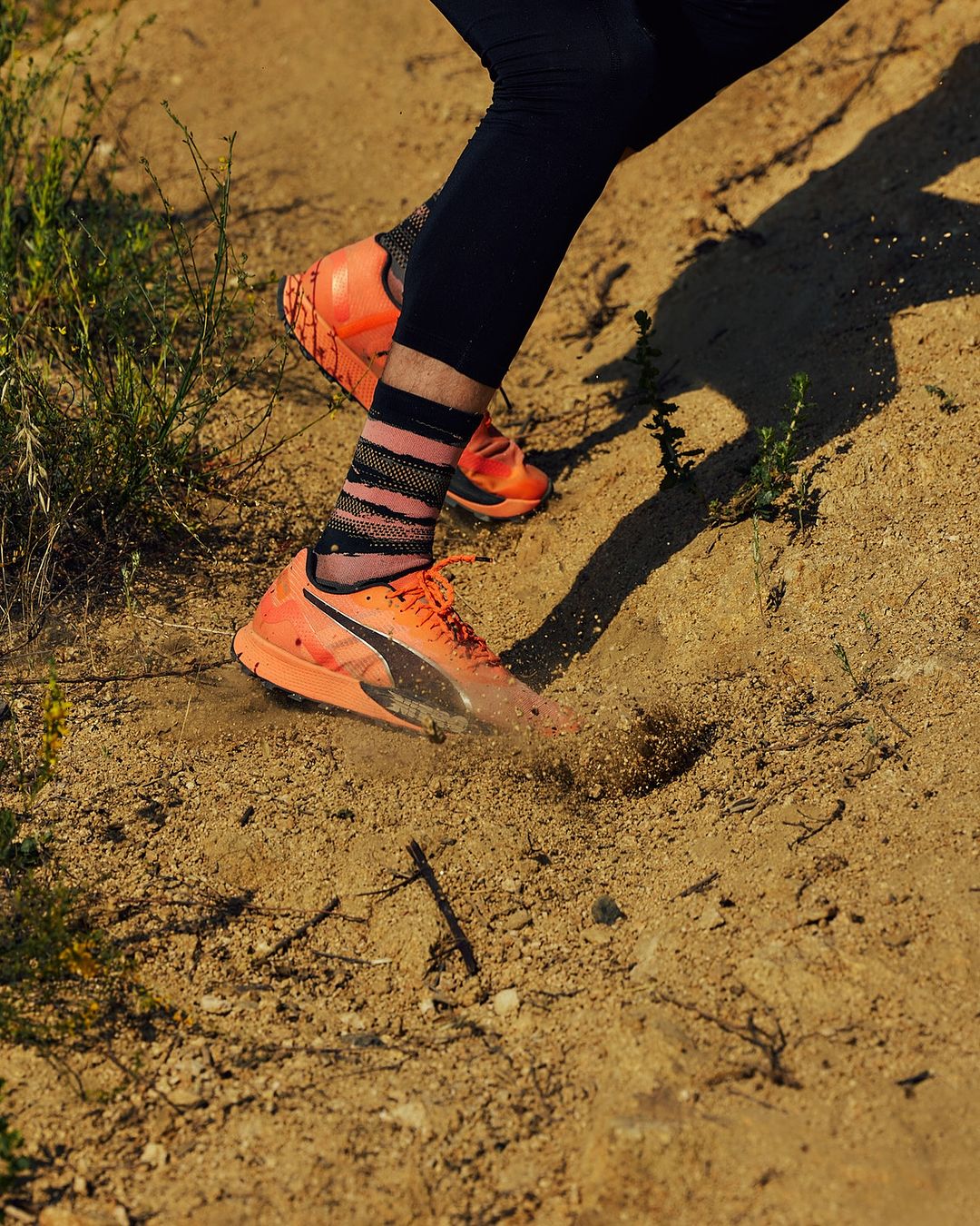 Close up of person running outdoors on sandy ground wearing orange Puma running shoes, black leggings and striped socks. Low angle.