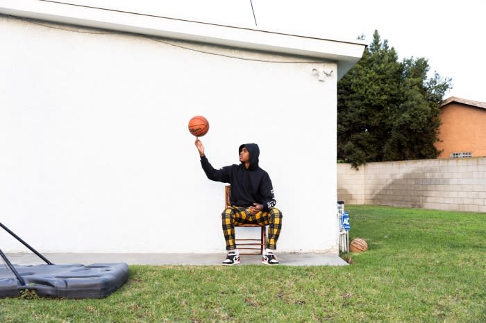 An African American male sits on a chair in a yard, spinning a basketball on one finger, wearing a dark hoodie and patterned pants.