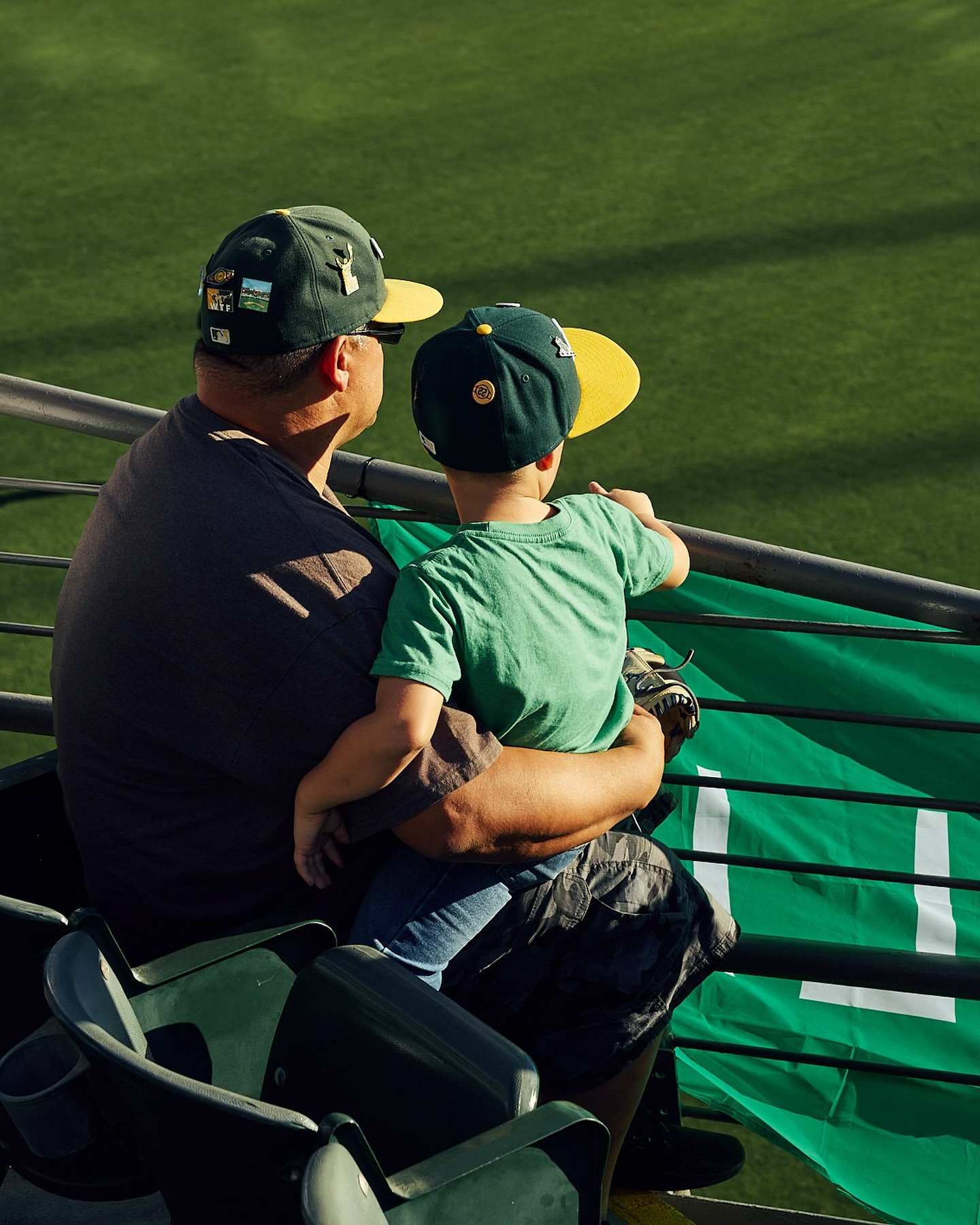 An adult and a child wearing baseball hats watch a game from stadium seats, featuring a green backdrop and a view from behind.