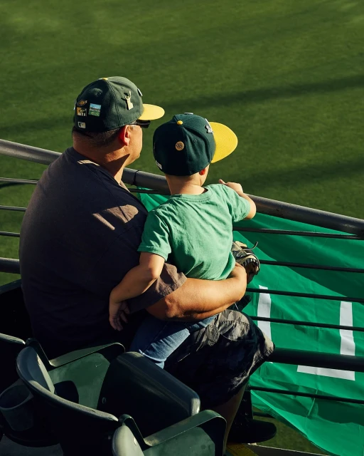 An adult and a child wearing baseball hats watch a game from stadium seats, featuring a green backdrop and a view from behind.