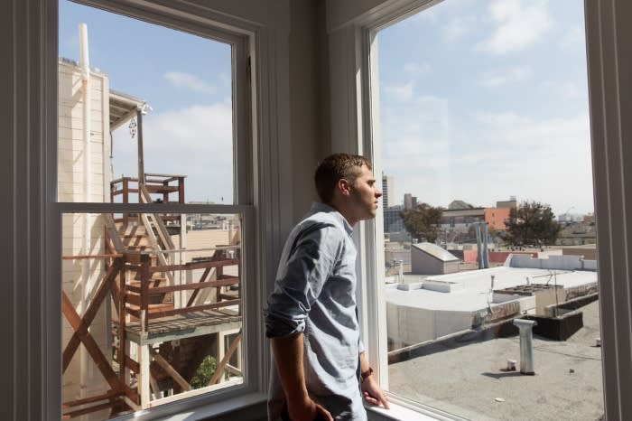Adult man in a button-down shirt gazes out of a window in an indoor setting, overlooking a cityscape and building under construction.