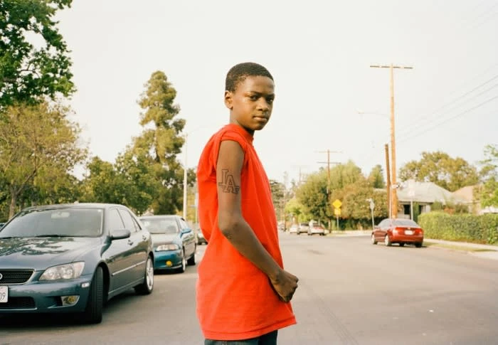 Young Black adult in a red t-shirt standing on an asphalt street in a residential area with cars and houses visible in the background.
