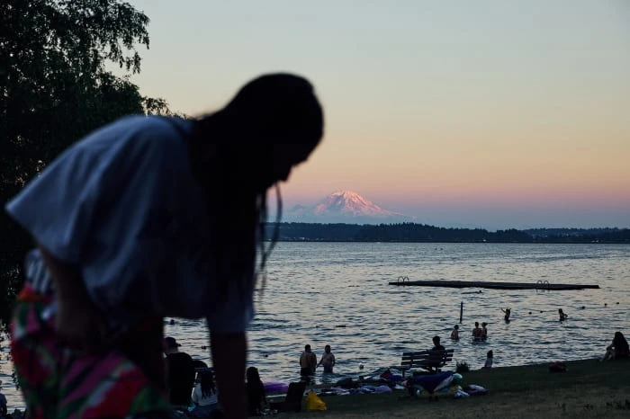 Scenic view of people swimming in a lake at sunset, with a snow-capped mountain in the distance and silhouetted trees in the foreground.