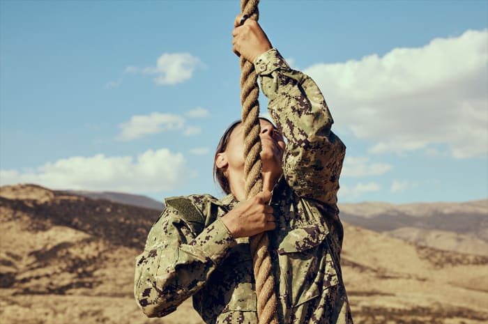 Adult female military person in camouflage uniform climbs a rope outdoors against a desert landscape and sunny sky with clouds.