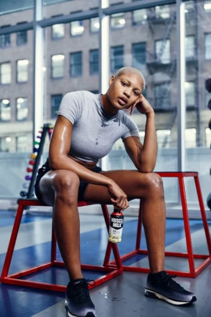 Athletic woman sitting in gym, holding Muscle Milk, wearing athletic top and shorts, with city view background, wellness concept.