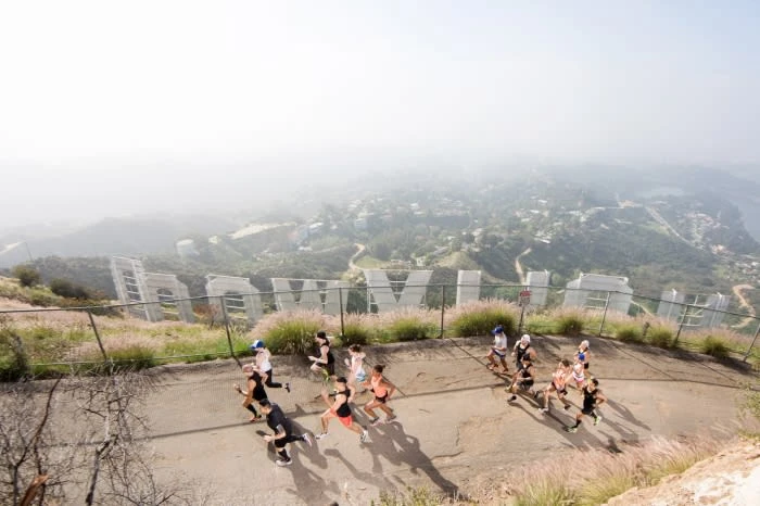 A group of people run uphill on a paved path, with the Hollywood sign and city visible in the background on an overcast day.