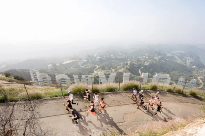 A group of people run uphill on a paved path, with the Hollywood sign and city visible in the background on an overcast day.