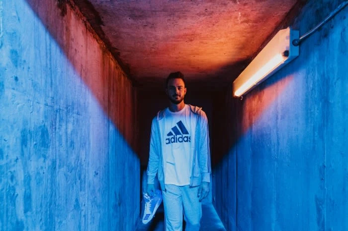Man in a hallway wears athletic brand apparel and holds a shoe. The concrete walls are lit with red and blue light.