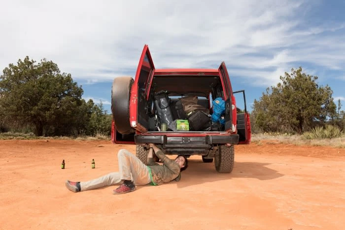 A man lying under a red SUV, working on the undercarriage, with the back doors open revealing bags, in a desert landscape with trees.