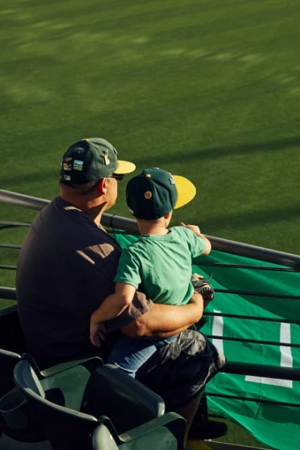 A father and son are watching a baseball game together while sitting in the stands; they both wear matching baseball caps.