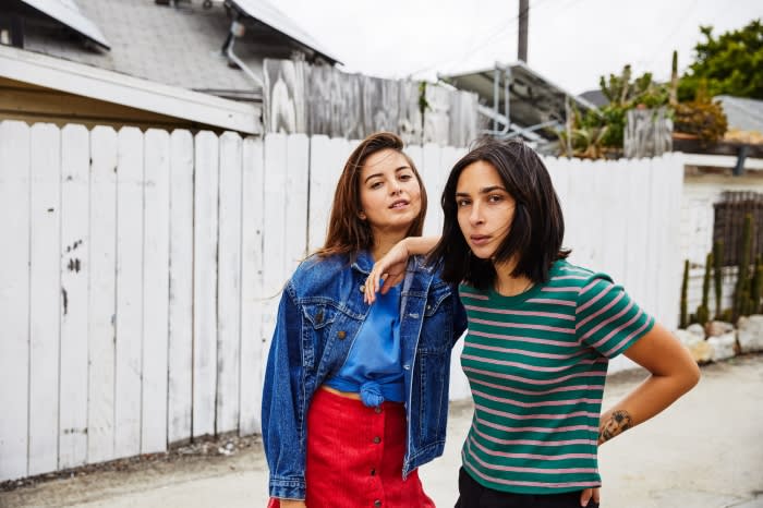 Two women stand close outdoors wearing fashionable outfits in a medium shot. One has a jean jacket and a red skirt, the other a striped shirt.