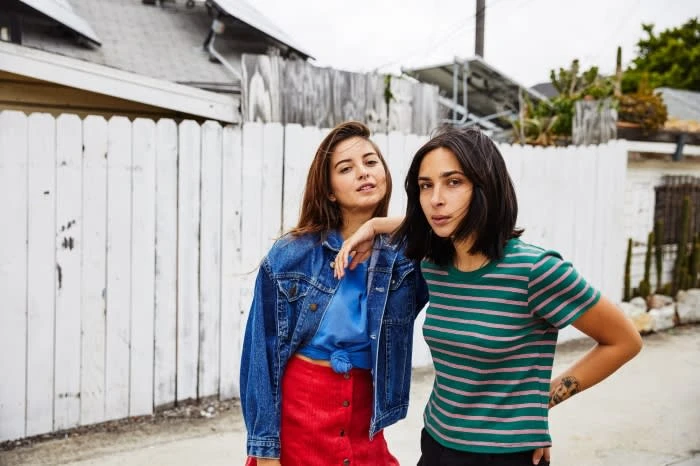 Two women stand close outdoors wearing fashionable outfits in a medium shot. One has a jean jacket and a red skirt, the other a striped shirt.