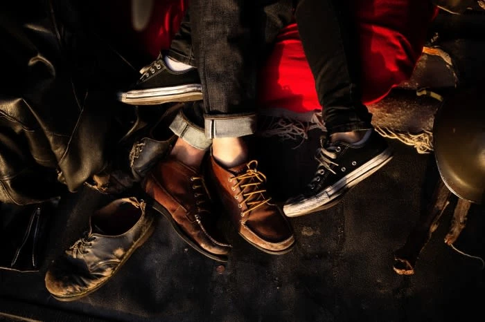 Close-up showcasing the lower bodies of two people wearing jeans and sneakers and dress shoes, sitting amidst leather jackets and luggage.