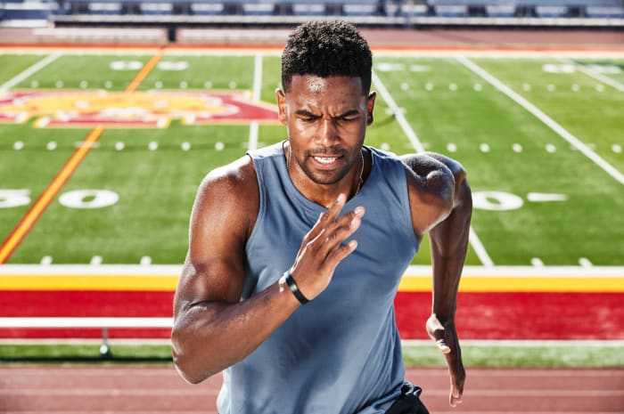 Athletic man runs at full speed in an outdoor stadium on a sunny day wearing a grey tank top and fitness tracker.