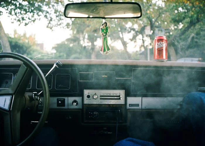 Vintage car interior featuring a steering wheel, dashboard with an analog radio, a Dr. Pepper can, and a hanging air freshener.