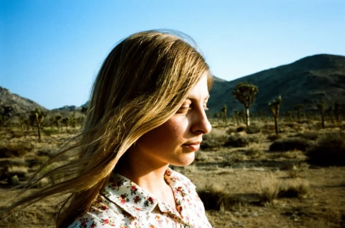 Profile portrait of a blonde woman wearing a floral shirt outdoors in a dry desert landscape with sparse trees and hills.