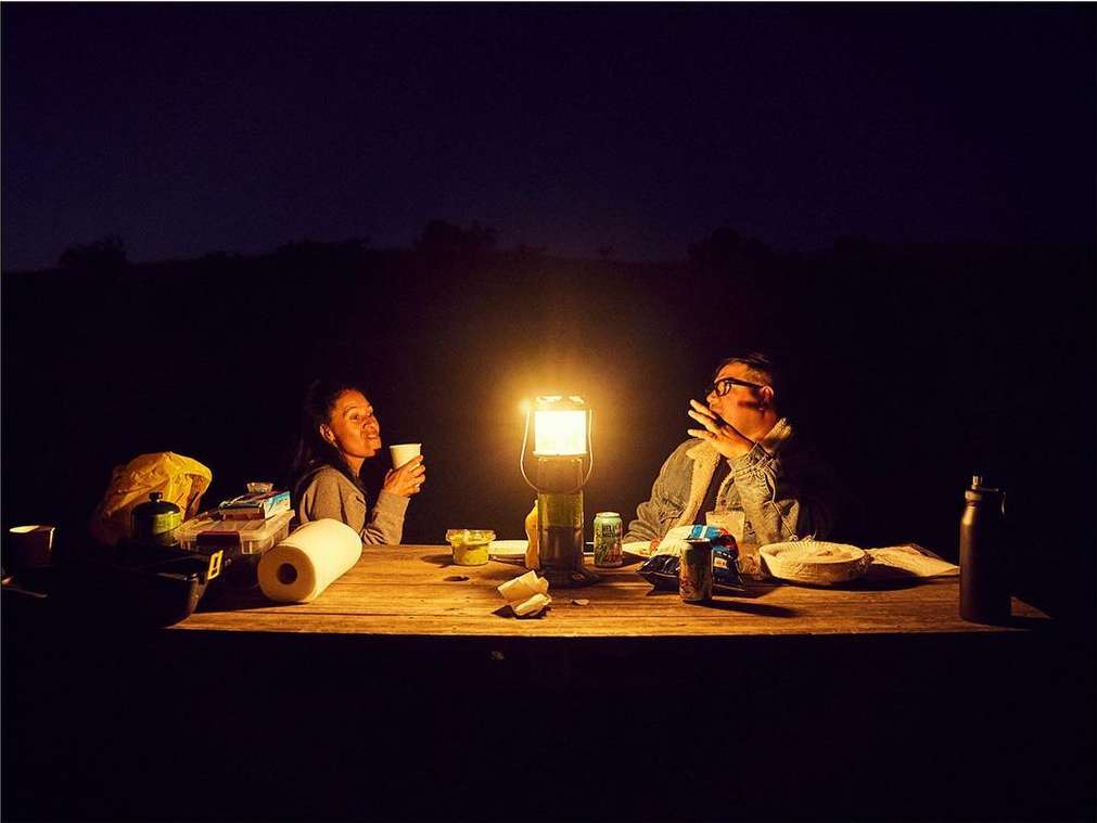 Two people sit at a wooden table illuminated by a lantern at night. The outdoor setting features food, drinks, and camping gear.