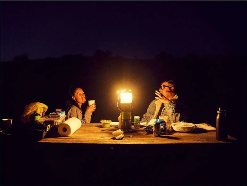 Two people sit at a wooden table illuminated by a lantern at night. The outdoor setting features food, drinks, and camping gear.