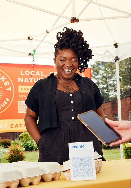 Smiling woman with styled dark hair stands at an outdoor market. She wears a dark romper and button-up shirt, taking payments via Venmo.