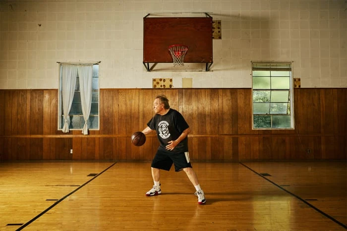 Man in black t-shirt dribbling basketball on wooden court inside gymnasium with basketball hoop, windows, and wood-paneled walls.