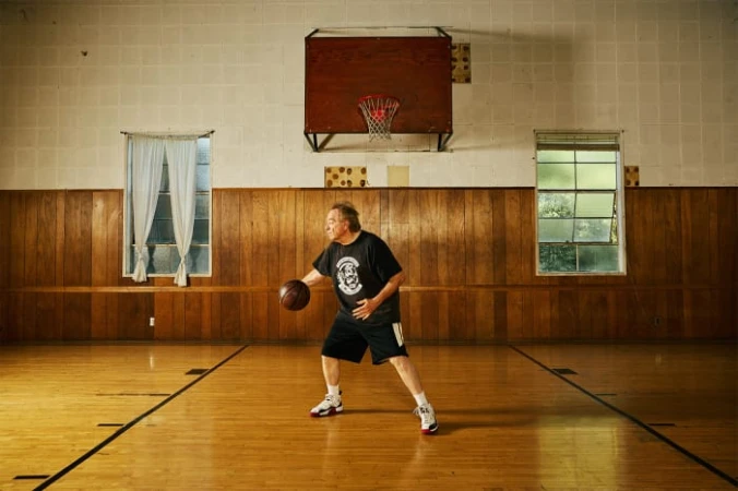 Man in black t-shirt dribbling basketball on wooden court inside gymnasium with basketball hoop, windows, and wood-paneled walls.