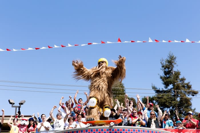 A large group of people watching a person in a Bigfoot costume on a skateboard, blue sky, and pennant banner overhead.
