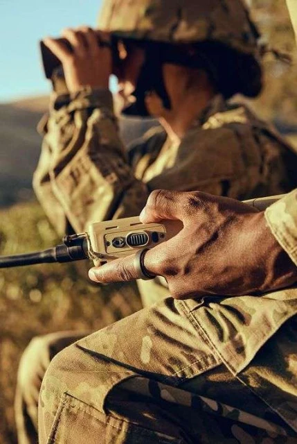 Two soldiers in camouflage uniforms, one using binoculars and the other holding a radio device, in an outdoor setting with natural light.